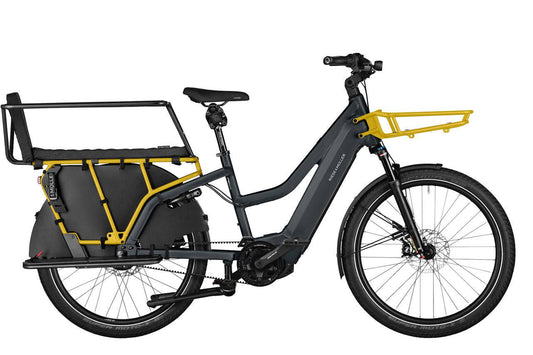 Ein schwarzes Riese & Müller Multicharger Mixte GT Fahrrad, mit gelben Vorderrad-Gepäckträger und schwarzen Gepäckträger hinten der mit einen gelben Gerüst an der Radnabe ünterstützend befestigt ist.